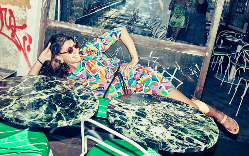 Smiling woman lies on a cafe table in a colourful dress and sunglasses, with chairs and tables in the background.