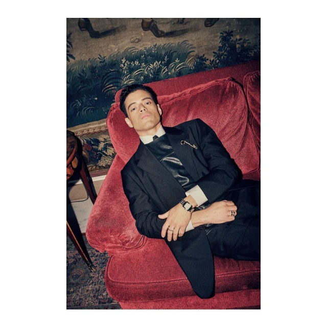 Man in a dark suit reclines on a red velvet couch in front of a vintage-style wallpapered wall, looking at the camera.