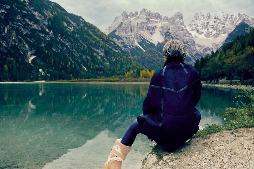 Adult female sits near a blue lake with snow-capped mountains in background; she wears a dark coat and has blonde hair and fair skin.