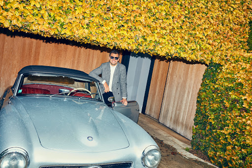 An adult male in sunglasses and blazer stands next to a silver vintage convertible car under a canopy of flowering yellow leaves.