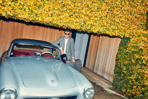 Man in grey blazer and sunglasses posing by a vintage silver convertible car under a vibrant yellow autumn foliage awning.