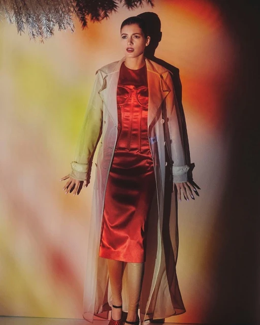 Woman poses in red dress and sheer coat against a colorful wall with soft lighting and a tree branch casting a shadow.