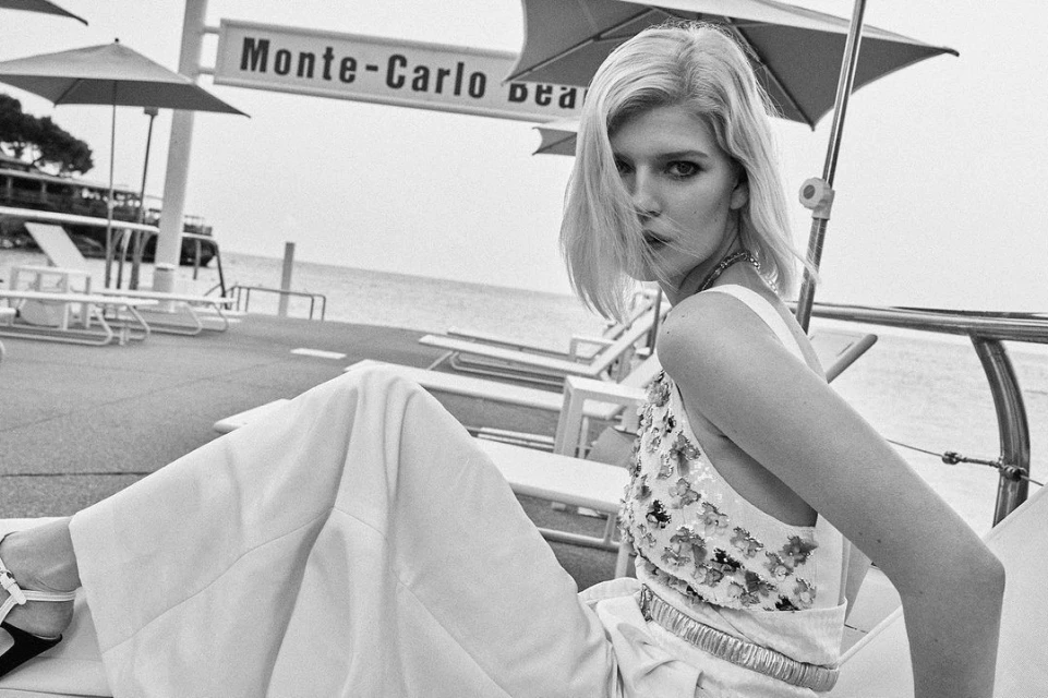 Monochrome fashion shot of a blonde adult female model posing on a beachside lounge chair with ocean and resort architecture in the background.