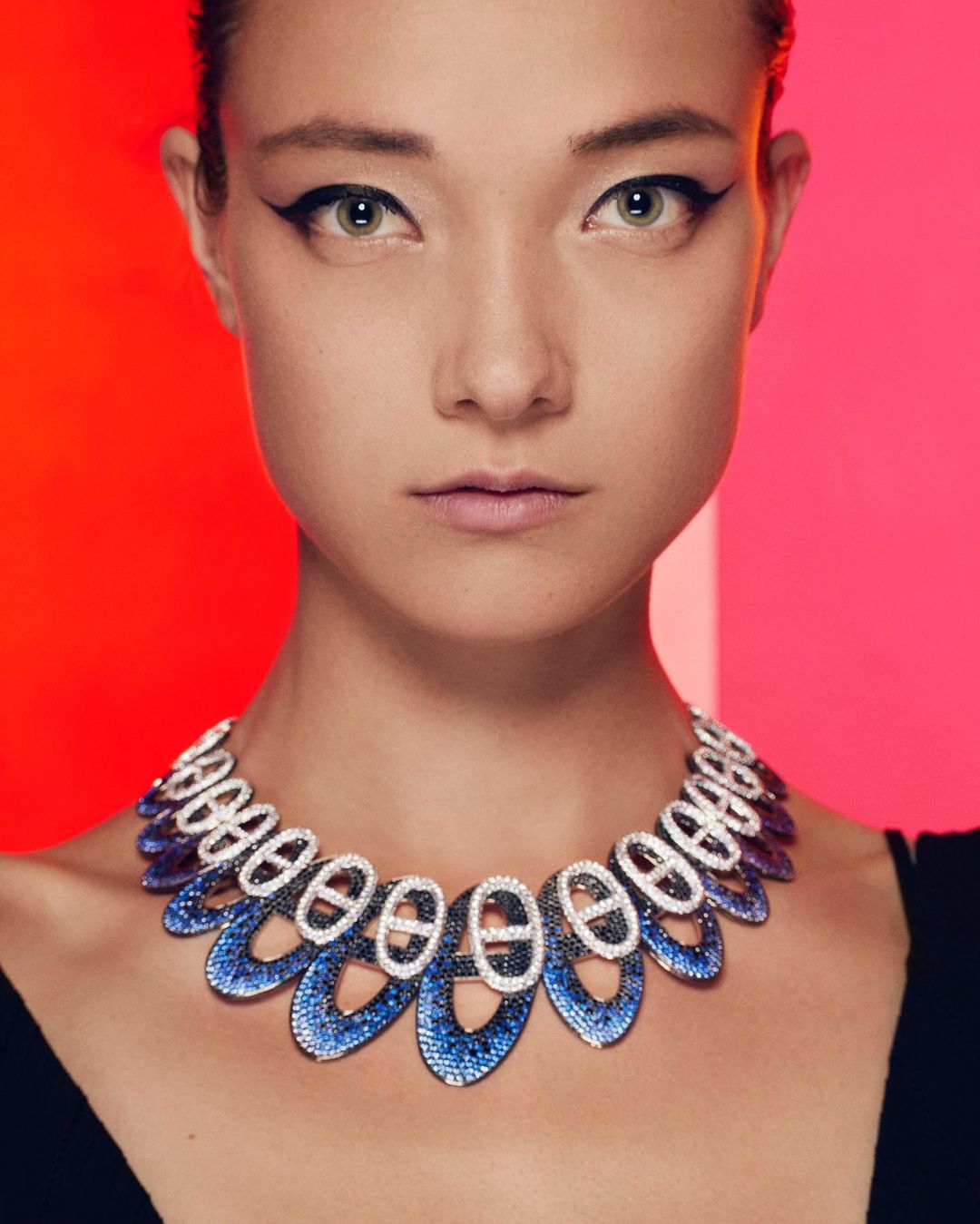 Close-up of an adult woman with dark eyeliner, wearing a blue and white decorative necklace against a red and pink background.