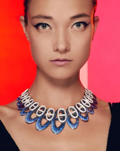 Close-up of an adult woman with dark eyeliner, wearing a blue and white decorative necklace against a red and pink background.