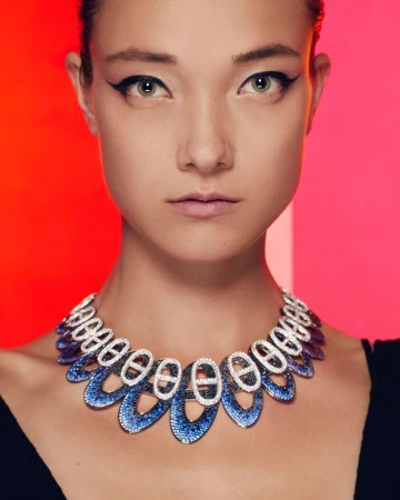Close-up of an adult woman with dark eyeliner, wearing a blue and white decorative necklace against a red and pink background.