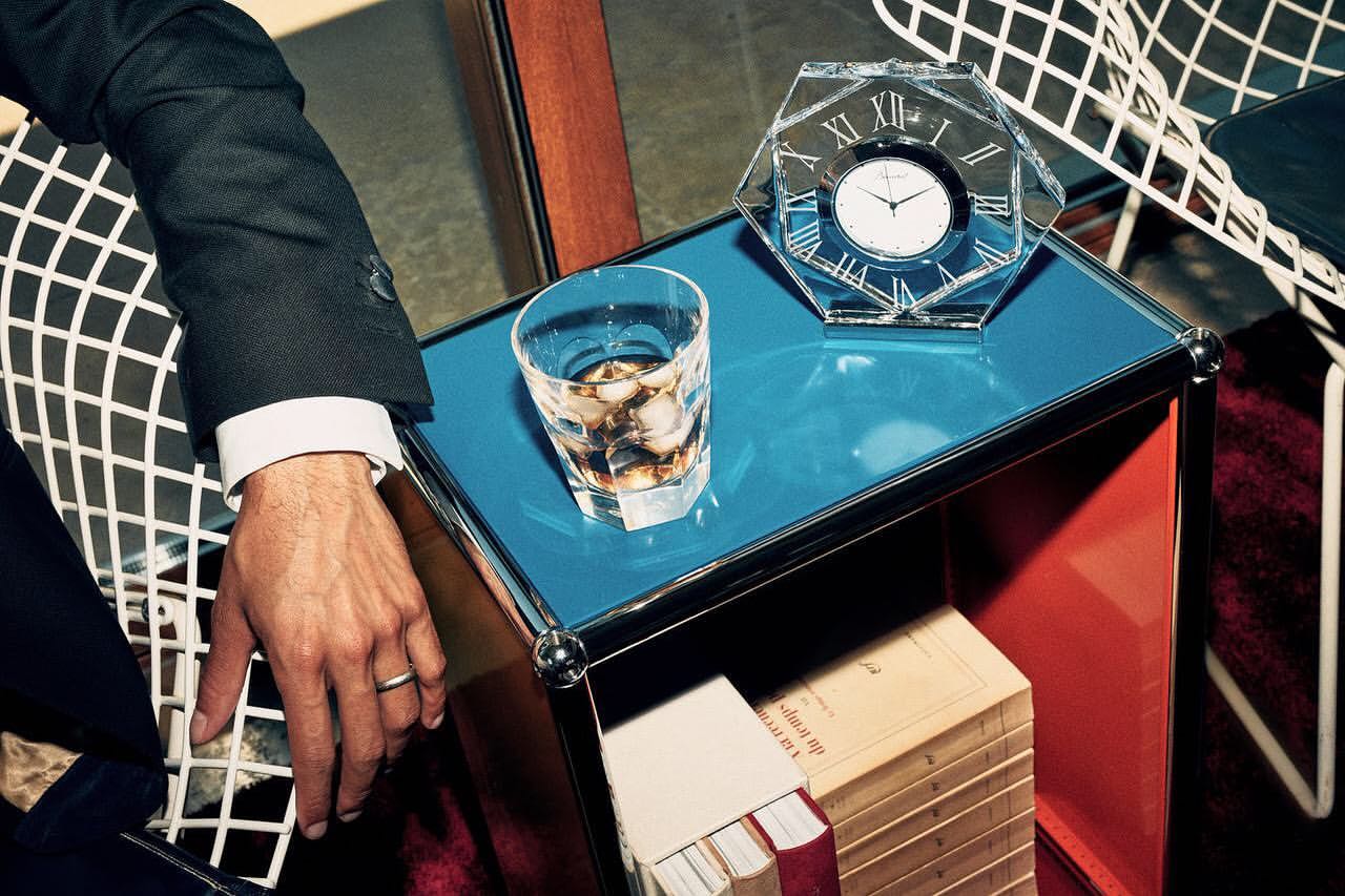 Close-up of a suited man's arm, a crystal glass of whiskey, and a crystal clock on a blue table alongside stacked books and a white chair.