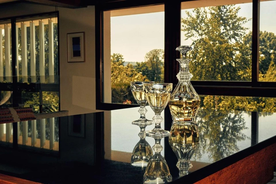 Crystal decanter and two glasses of white wine on a dark reflective surface in front of a large window overlooking greenery.