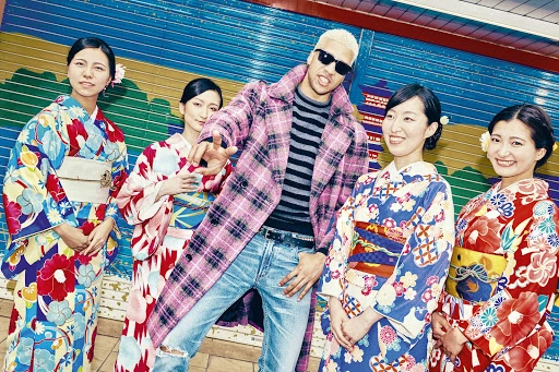 Group of women in kimonos pose with a man in sunglasses and a pink plaid coat against a blue and yellow painted backdrop.