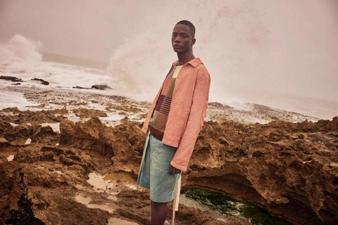 Black man standing on a rocky shoreline wearing a pink jacket and light blue shorts as ocean waves crash in the background.