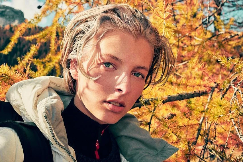 Close-up portrait of a young adult with blond hair and a light complexion, wearing a jacket, set against a backdrop of autumn foliage.