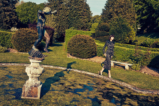 Woman in long-sleeved patterned dress walks through a lush green garden with manicured hedges, trees, and a statue fountain feature.