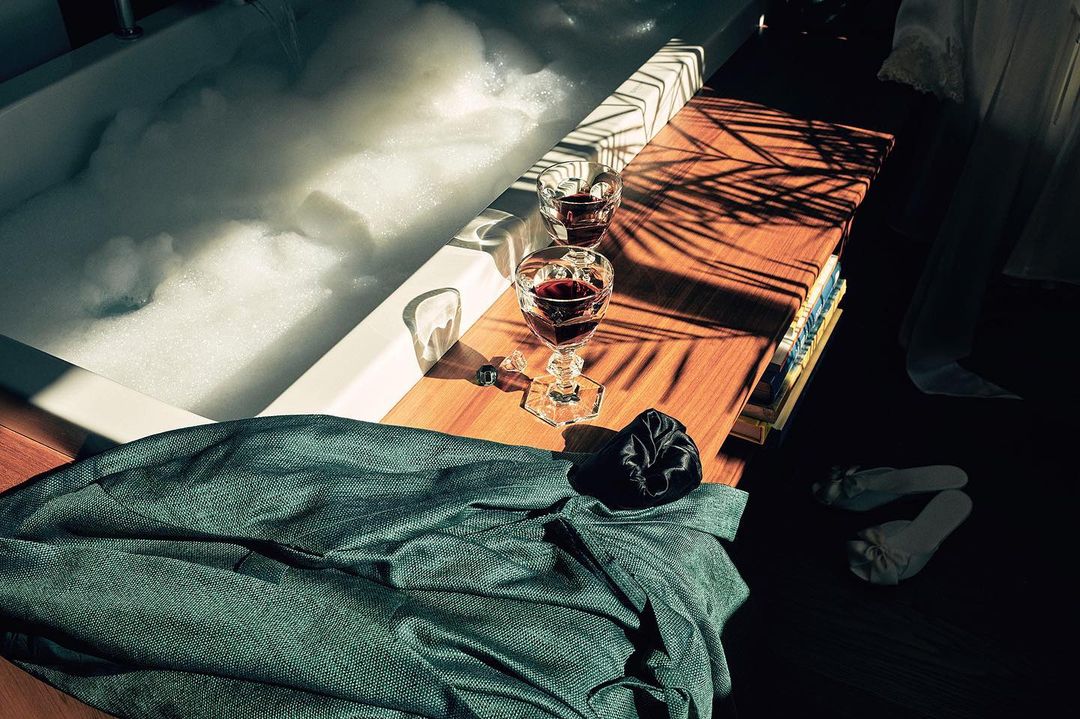 Bathtub filled with bubbly water, wine glasses on a wooden surface, and a green dress on a wooden floor.