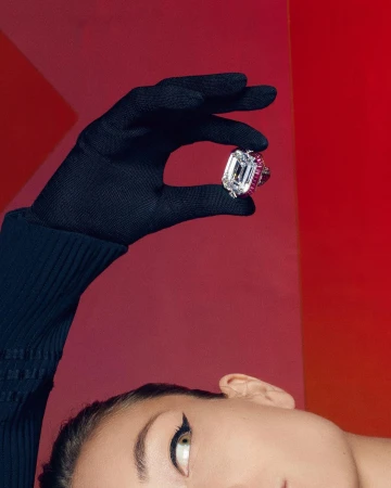 Close-up of a person in a black glove holding a large emerald-cut diamond ring, with their eye and winged eyeliner visible below.