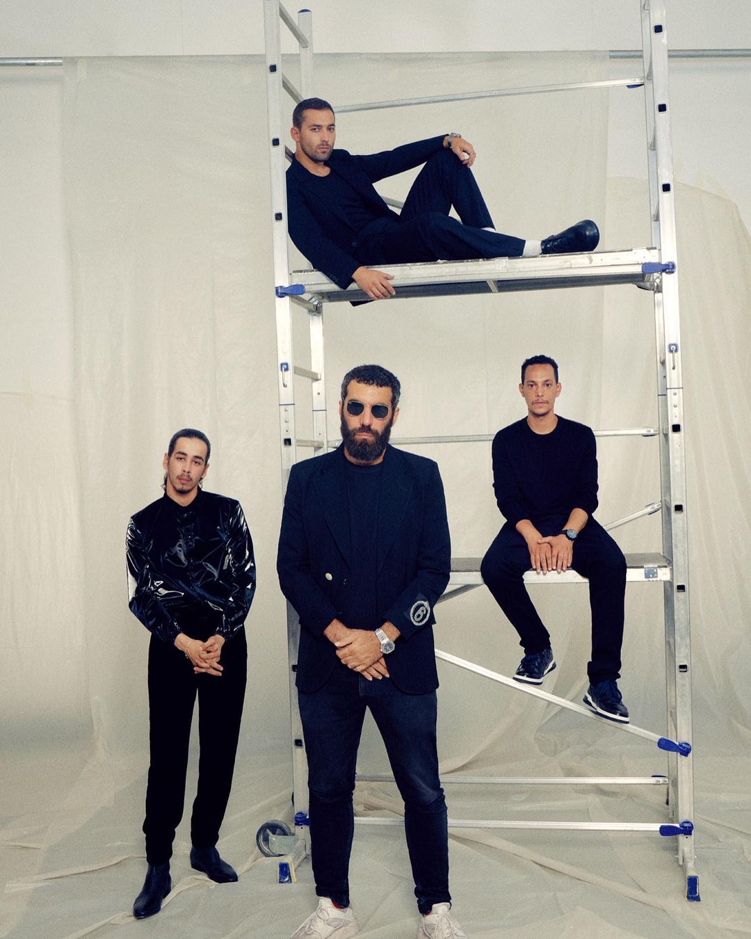 Four men in fashionable all-black attire pose near a silver aluminum step ladder in a minimalist indoor setting for a portrait.