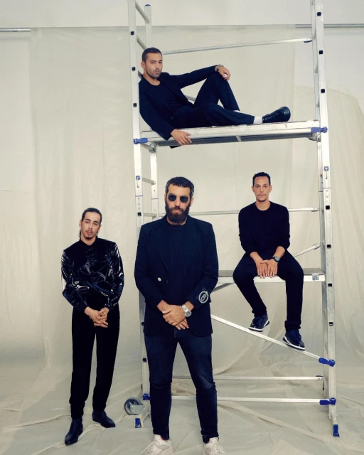 Four men in fashionable all-black attire pose near a silver aluminum step ladder in a minimalist indoor setting for a portrait.