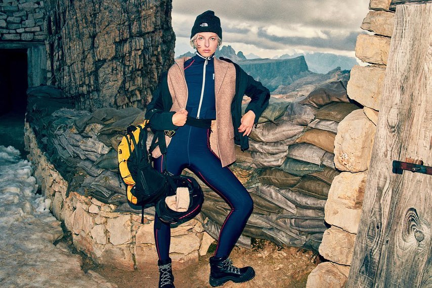Woman in outdoor apparel stands in a stone structure with mountains in the background, wearing a beanie, jacket, and hiking boots.