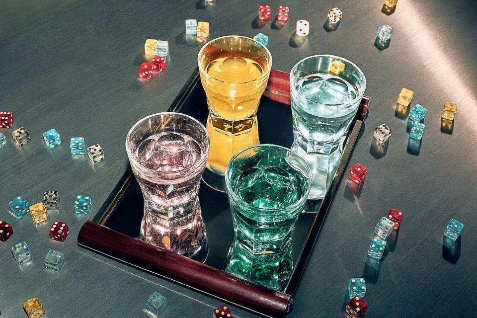 Four glasses filled with different colored liquids sit on a mirrored tray amidst scattered dice on a textured grey surface.