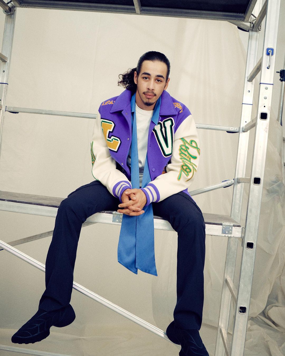 Stylish man sits on aluminum ladder in photo studio wearing purple and off-white varsity jacket, blue scarf, and black athletic shoes.