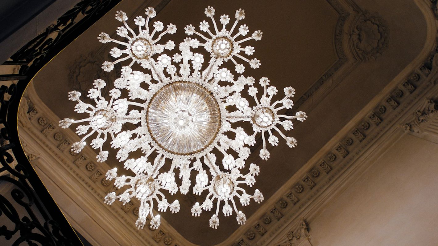Ornate crystal chandelier hangs from a ceiling with decorative moulding in an indoor setting, creating an elegant, vintage ambiance.