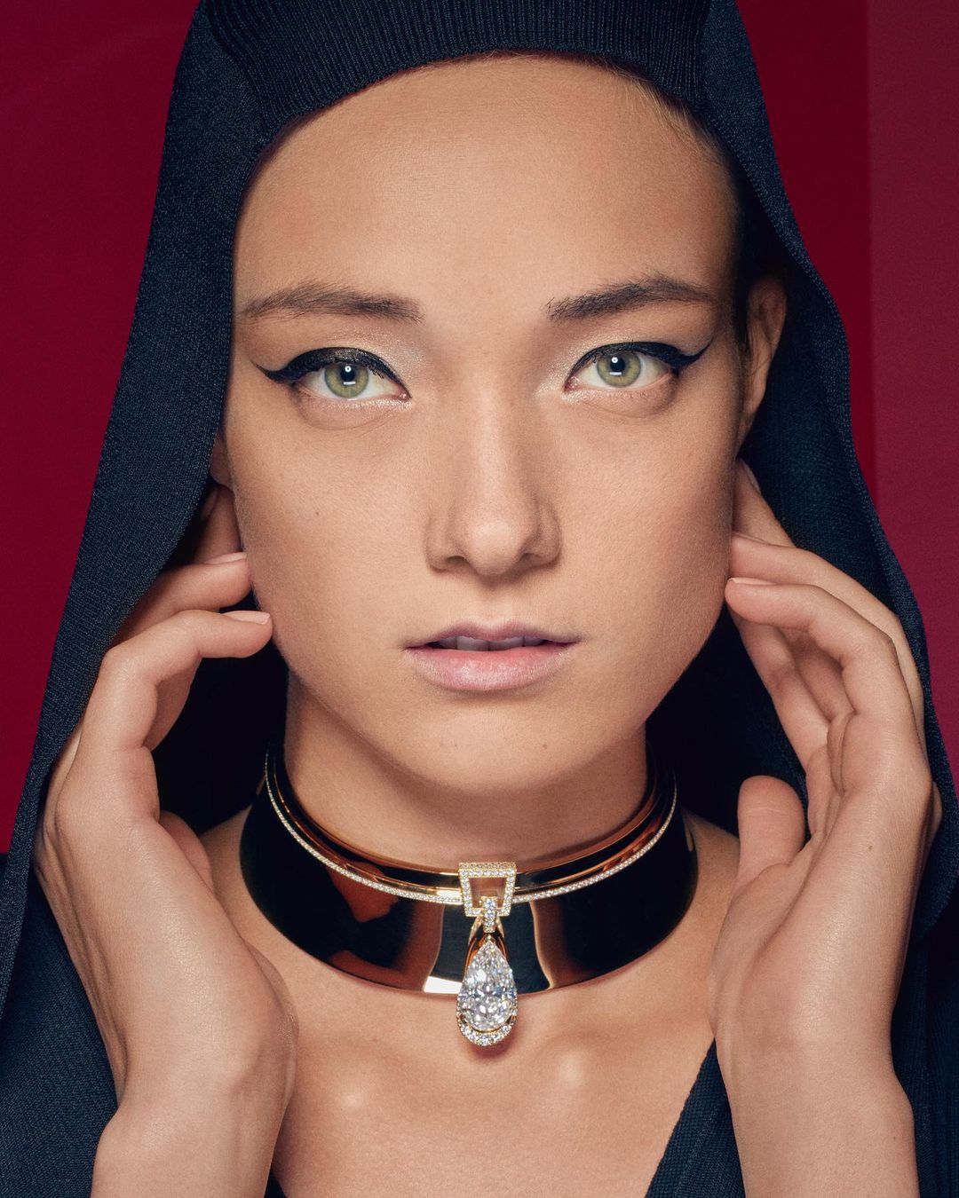 Glamorous portrait of a woman with bold eyeliner wearing an elegant gemstone collar necklace against a deep red backdrop.