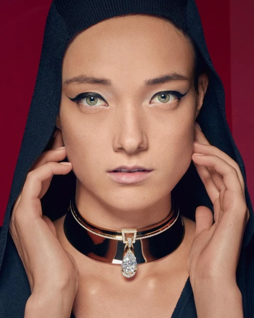 Glamorous portrait of a woman with bold eyeliner wearing an elegant gemstone collar necklace against a deep red backdrop.