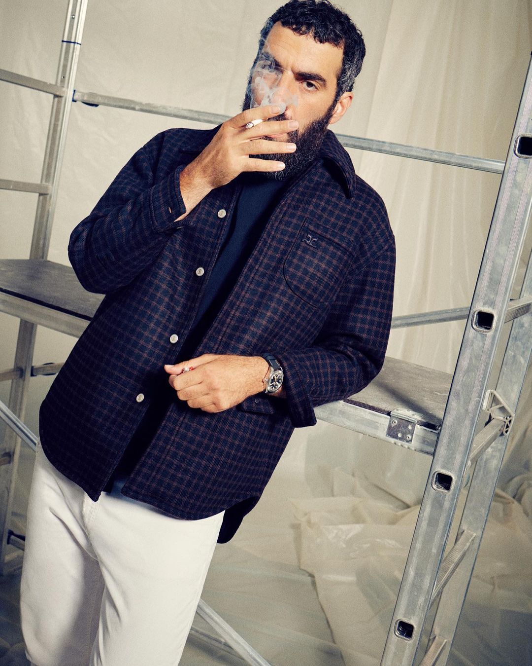 Adult man with beard in checkered jacket smoking cigarette next to aluminum ladder in studio with white plastic sheeting backdrop.