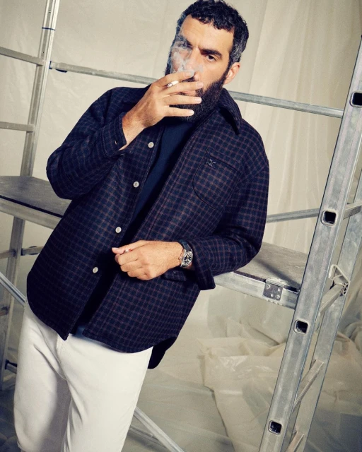 Adult man with beard in checkered jacket smoking cigarette next to aluminum ladder in studio with white plastic sheeting backdrop.