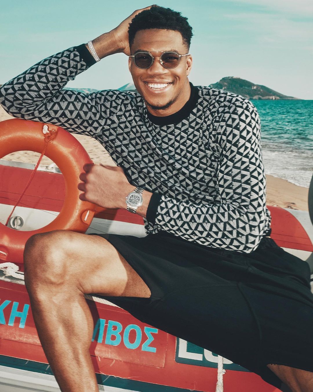 Smiling adult male athlete in patterned sweater and sunglasses seated near a red life preserver against a beach and sky backdrop.