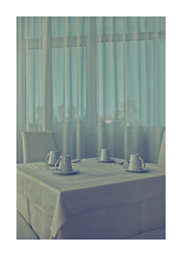 Four white ceramic tea cups sit on saucers atop a square white tablecloth on a table in a brightly lit room with white curtains.