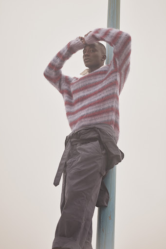 Adult male wearing striped sweater poses leaning against pole against a plain background in an outdoor setting with natural lighting.