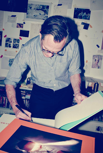 Adult male in glasses signs book in indoor setting with desk, papers, and book shelves as background. Art and editorial elements.