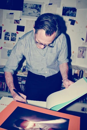 Adult male in glasses signs book in indoor setting with desk, papers, and book shelves as background. Art and editorial elements.