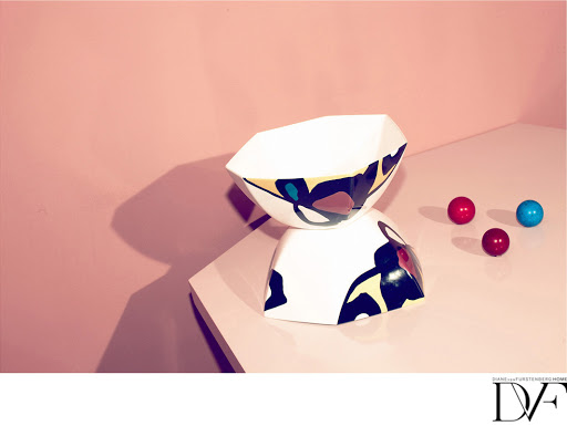 Studio shot of two geometric, patterned ceramic bowls stacked, alongside three colored marbles, set against a pink wall backdrop.