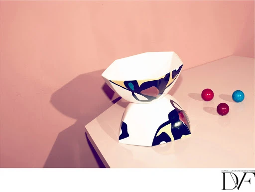 Studio shot of two geometric, patterned ceramic bowls stacked, alongside three colored marbles, set against a pink wall backdrop.