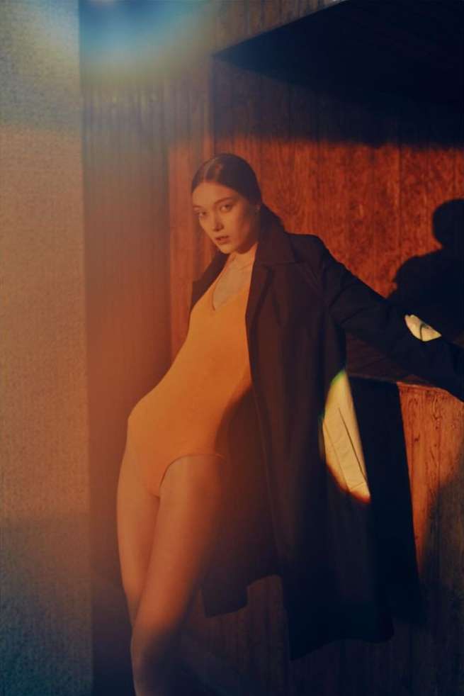 Indoor portrait of an adult woman in yellow bodysuit with dark coat leaning against a wall with mixed lighting, studio setting.