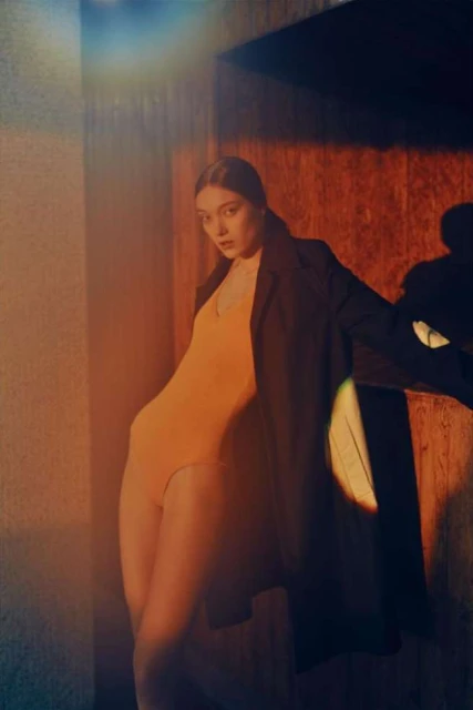 Indoor portrait of an adult woman in yellow bodysuit with dark coat leaning against a wall with mixed lighting, studio setting.