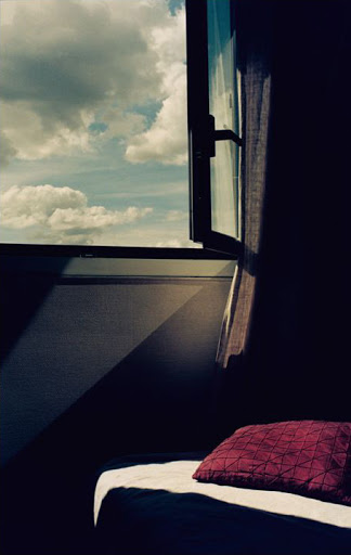 Open window view reveals a sky filled with fluffy clouds, casting a beam of light on a pillow in a dimly lit room interior.