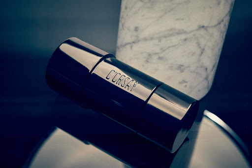 A cylindrical, dark metallic container with 'C'ORSAY' engraved in all caps, rests on a glossy surface against a marble backdrop.