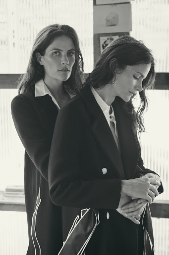 Two adult women pose indoors wearing dark blazers and collared shirts, one with her arms around the other from behind in a medium shot.