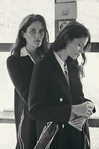 Two adult women pose indoors wearing dark blazers and collared shirts, one with her arms around the other from behind in a medium shot.