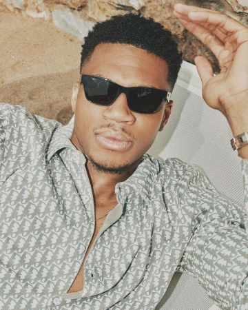 Portrait of a Black man wearing dark sunglasses and a patterned grey button-down shirt relaxing outdoors