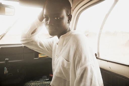 Close-up of a South Sudanese adult female wearing a white shirt, sitting in a vehicle looking at the camera with a neutral expression.