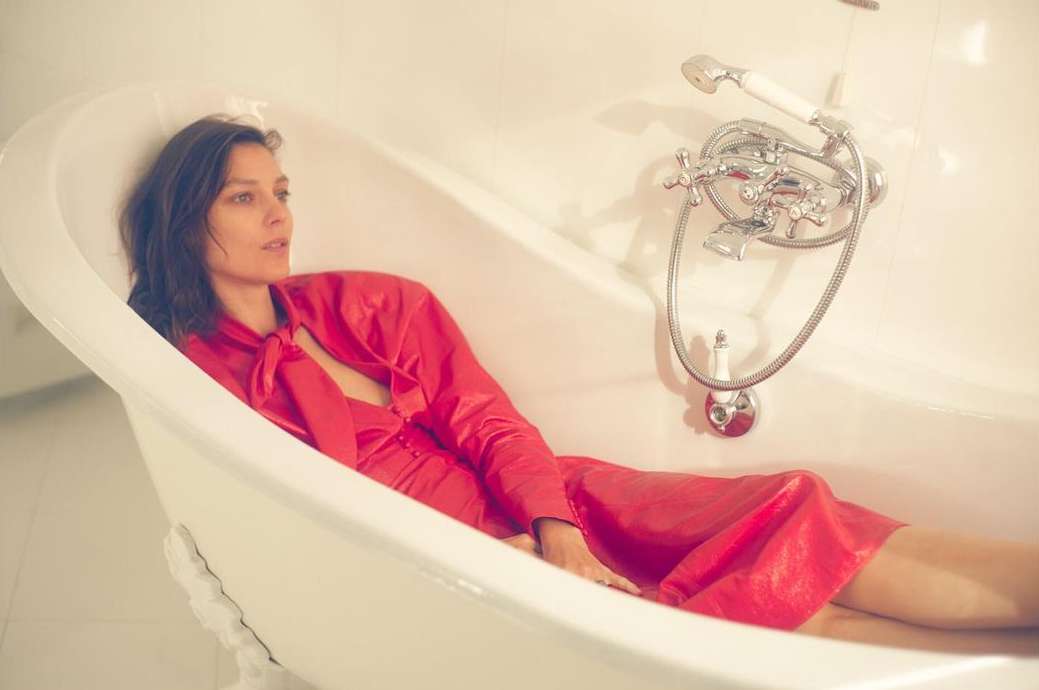 A woman in a red dress reclines in a white clawfoot bathtub, her relaxed pose complemented by the neutral bathroom setting.