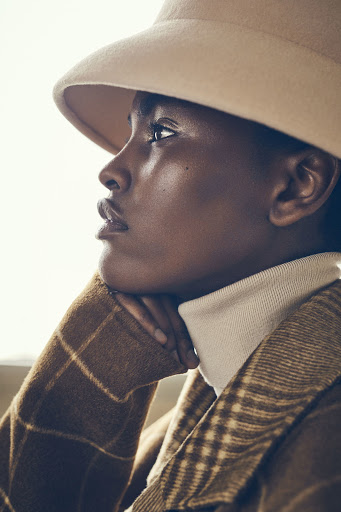 Profile shot of a Black woman wearing a beige bucket hat and a brown and tan plaid jacket with a beige turtleneck sweater.
