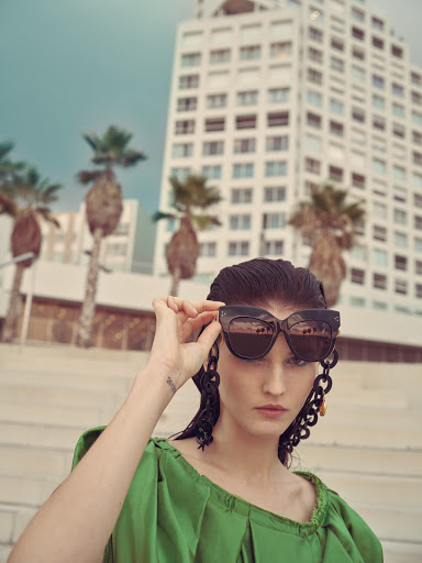 Elegant woman in green dress and sunglasses poses outdoors with modern architecture and palm trees in soft, natural light for a fashion editorial.