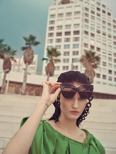 Elegant woman in green dress and sunglasses poses outdoors with modern architecture and palm trees in soft, natural light for a fashion editorial.