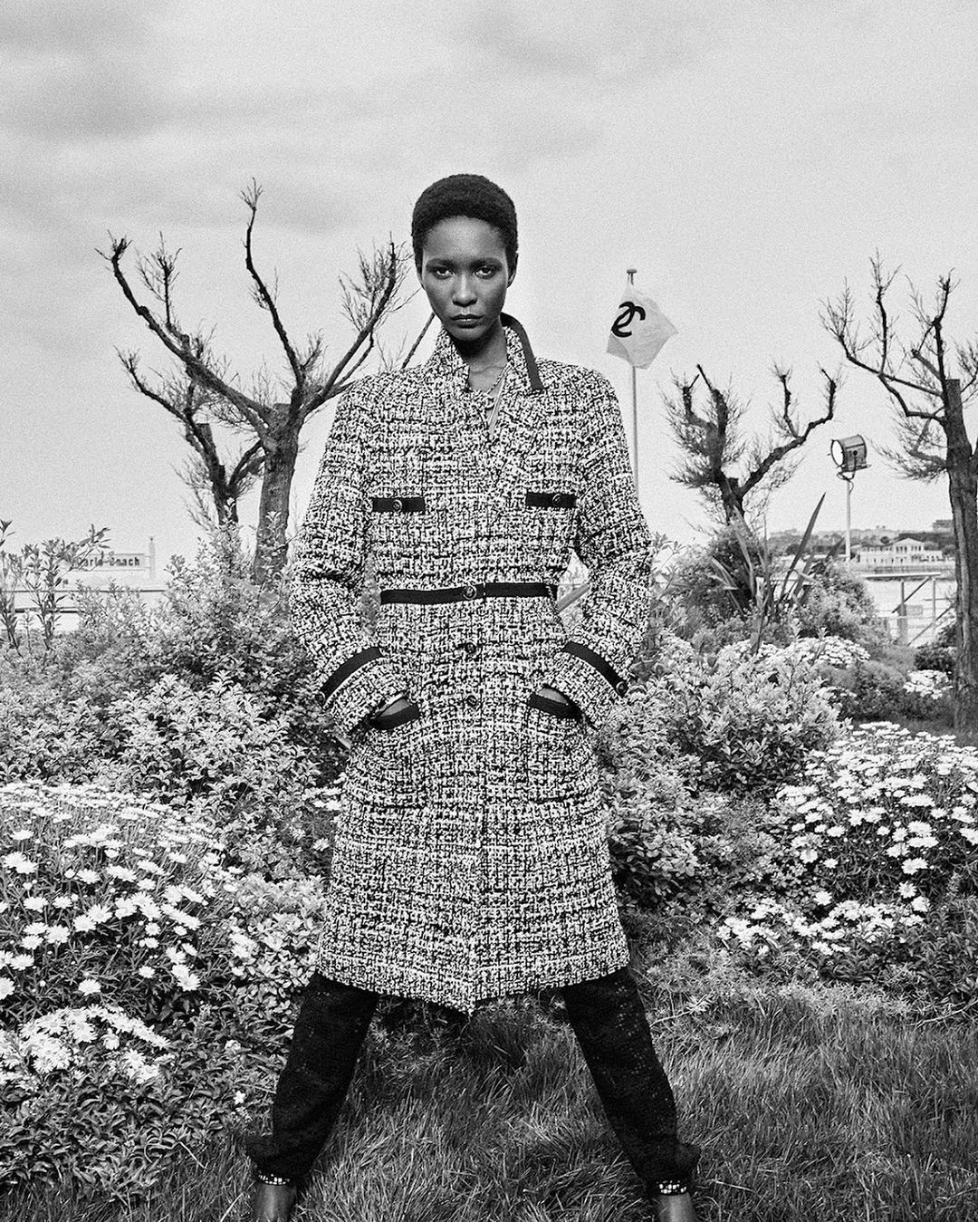 Black and white portrait of a Black female model in a tweed coat, standing in a park with bare trees and wildflowers, fashion shot.