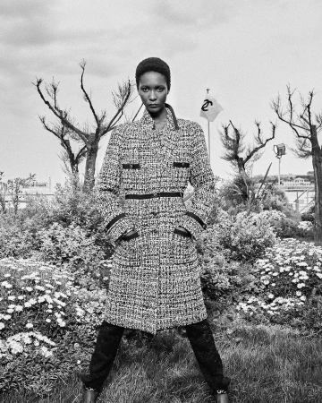 Black and white portrait of a Black female model in a tweed coat, standing in a park with bare trees and wildflowers, fashion shot.