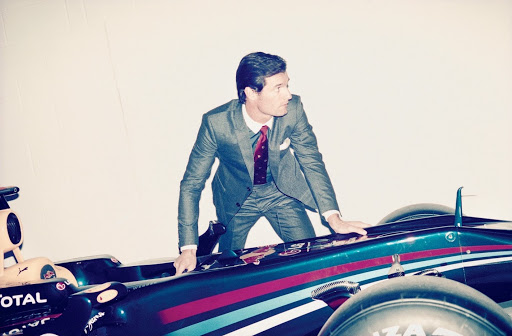 Man in a suit leans on a green and red Formula 1 race car in a photo studio, posing for a fashion editorial. 